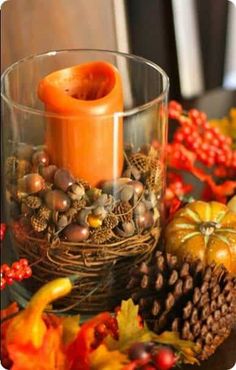 an orange candle is sitting in a glass bowl with pine cones and acorns