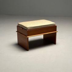 a small wooden bench with a cushion on it's top and bottom shelf, in front of a gray background