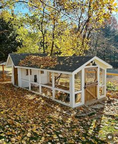 a white chicken coop in the middle of leaves on the ground and trees with fall foliage around it