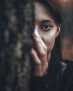 a woman with blue eyes is looking out from behind a tree trunk and holding her hand to her face