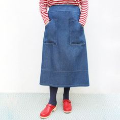 a woman standing in front of a white wall wearing a blue denim skirt and red shoes