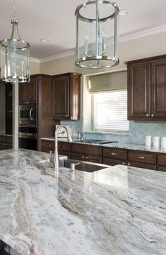 a large kitchen with marble counter tops and wooden cabinets, along with two hanging lights