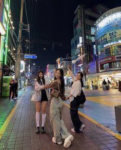 three girls are standing on the sidewalk in front of some buildings at night, and one girl is holding her hand up