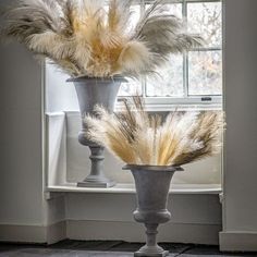 two vases with feathers on them in front of a window, one is gray and the other is white