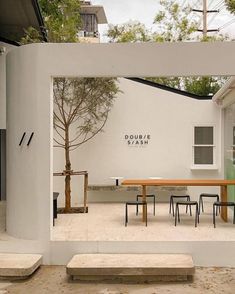 a table and benches in front of a white building with a tree on the other side