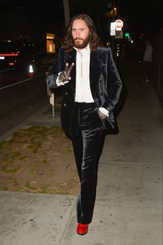 a man with long hair wearing a suit and red shoes walking down the street at night