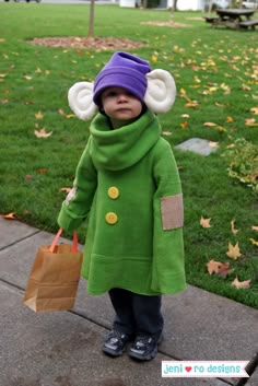 a little boy in a green coat and purple hat holding a brown paper bag while standing on the sidewalk