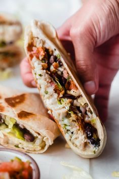 a person holding a burrito in their hand with other food on the table behind it