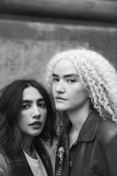 two women standing next to each other in front of a stone wall with long curly hair