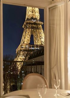 the eiffel tower is lit up at night from inside a restaurant window with white tables and chairs