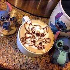 a cup of coffee with an image of stitch and stitch on it next to two small figurines