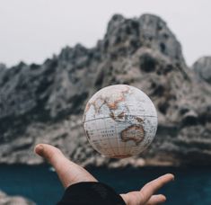 a person holding up a small globe in front of a mountain and body of water