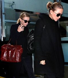 two women in black coats and sunglasses are walking down the street with their luggage behind them