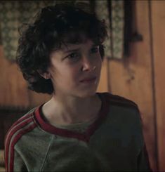 a young boy with curly hair standing in front of a wooden wall and looking at the camera