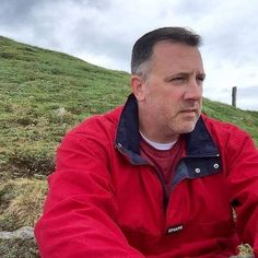 a man in a red jacket sitting on top of a grass covered hill next to a fence