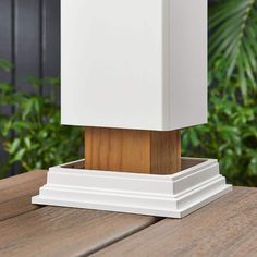 a white lamp sitting on top of a wooden table in front of some bushes and trees