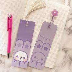 two purple bookmarks sitting on top of a table next to a pink pen