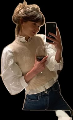 a woman holding a wine glass and looking at her cell phone while wearing a sweater