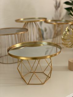 three round tables with gold metal bases and glass tops on a white countertop next to plants