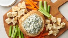 a wooden cutting board topped with bread and veggies next to a bowl of dip