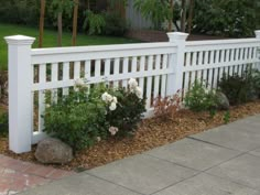 a white picket fence with flowers and rocks in the foreground on a brick walkway