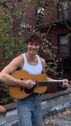 a young man holding an acoustic guitar in front of a brick building and smiling at the camera