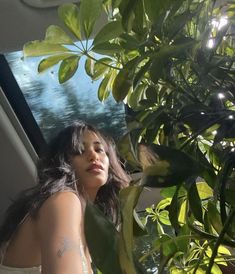 a woman in a white dress standing next to a green plant and looking out the window