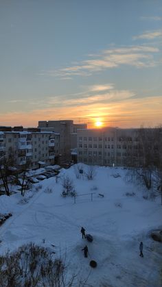 the sun is setting over some buildings and people are walking in the snow on the ground