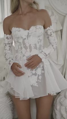 a woman wearing a white dress with flowers on the sleeves and skirt is posing in front of a bed