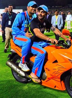 two men sitting on an orange motorcycle in the middle of a soccer field at night