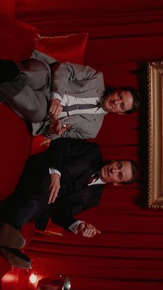 two men dressed in suits and ties posing for a photo on a red couch with a gold framed mirror behind them