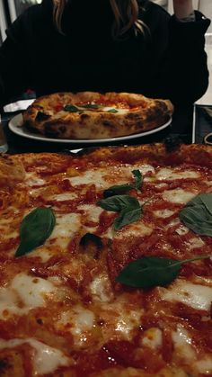 a woman sitting at a table in front of two pizzas with basil leaves on them