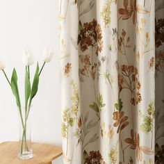 two white tulips in a vase on a table next to a floral curtain