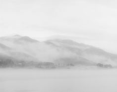 a black and white photo of mountains in the fog on a lake with water below