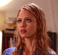 a woman with long blonde hair and blue shirt looking at the camera while standing in front of a bookshelf