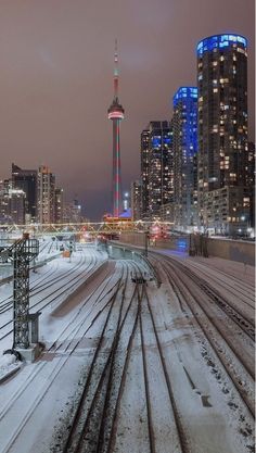 the city skyline is lit up at night with snow on the ground and tracks running through it