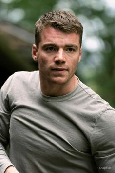 a man in grey shirt looking at the camera with trees in the back ground behind him