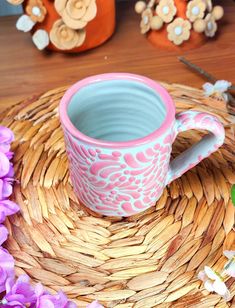 a pink and blue coffee cup sitting on top of a wicker place mat next to purple flowers