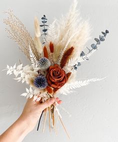 a hand holding a dried flower bouquet with feathers and flowers in it's center