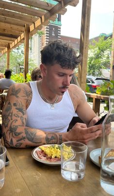 a man with tattoos sitting at a table looking at his cell phone and eating food