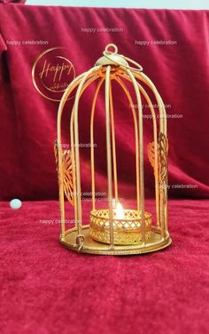 a small gold birdcage with a candle inside on a red velvet surface next to a pillow