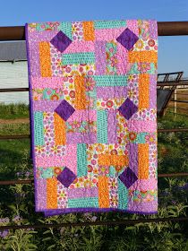 a patchwork quilt hanging on a fence in front of some flowers and a barn