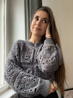 a woman wearing a gray crochet sweater looks out the window while holding her hand on her head