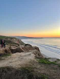 three people standing on the edge of a cliff overlooking the ocean and beach at sunset