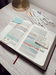 an open book sitting on top of a wooden table next to pens and a lamp