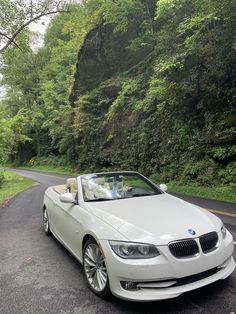 a white bmw convertible parked on the side of a road in front of a lush green forest