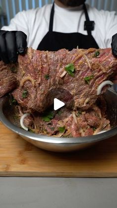 a man in an apron holding up a large piece of meat with onions and herbs on it