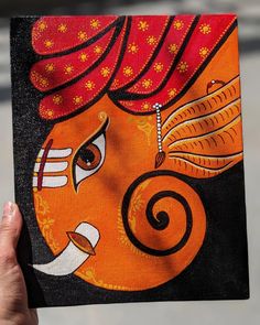 a hand holding up an orange and black painting on canvas with the face of ganesh