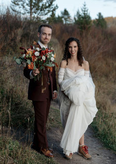 a man and woman walking down a dirt road holding flowers in their hands while dressed in wedding attire