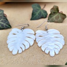 two white leaf shaped earrings sitting on top of a wooden table next to green leaves